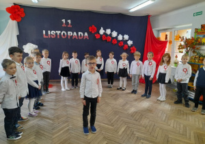 Dzieci z grupy "Słoneczka" stoją ubrane w stroje galowe z przypiętymi kotylionami w barwach narodowych. Na środku sceny Mikołaj zaprasza przybyłych gości na przedstawienie. W tle dekoracja - do granatowego płótna przypięty jest napis 11 Listopada oraz białe i czerwone kwiaty. Po bokach wisi białe i czerwone płótno.