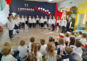 Dzieci stoją w półkolu na tle dekoracji i śpiewają piosenkę pt. "11 Listopada". Przedszkolaki z innych grup siedzą na dywanie i oglądają występ kolegów.