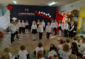 Bartek, Mikołaj i Tosia stoją na środku sceny deklamują wiersz pt. "Mały patriota", a za nimi stoją rówieśnicy. W tle dekoracja z okazji Święta Niepodległości. Młodsi koledzy siedzą na dywanie i oglądają przedstawienie.