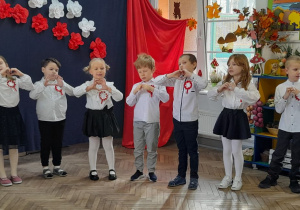 Siedmioro dzieci stoi obok siebie podczas występu i śpiewa piosenkę patriotyczną tworząc serduszka z rączek.