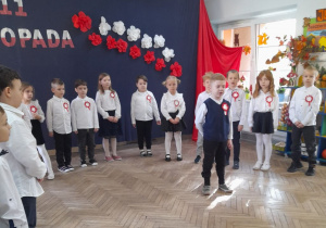 Oliwier stoi na środku sceny i recytuje fragment wiersza o Polsce. Za chłopcem stoją rówieśnicy. W tle dekoracja z okazji Święta Niepodległości.