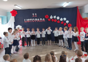 Dzieci stoją w półkolu na tle dekoracji i śpiewają "Hymn małego patrioty". Przedszkolaki tworzą z rączek serduszka.