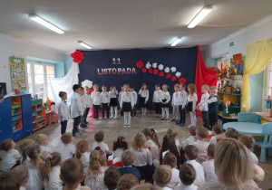 Przedszkolaki siedzą na dywanie i oglądają występ z okazji Święta Niepodległości. Marysia stoi na środku sceny i recytuje kolejny fragment wiersza o Polsce, a za nią stoją rówieśnicy. W tle dekoracja.