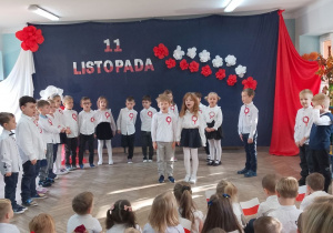 "Słoneczka" stoją w półkolu na tle dekoracji, a Bruno i Emma stoją na środku sceny i recytują fragment wiersza o Polsce.