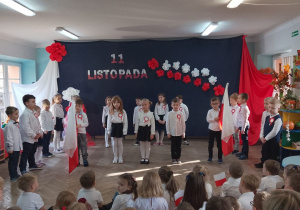 Pięcioro dzieci stoi na środku sceny, a za nimi rówieśnicy. Dwóch chłopców trzyma biało-czerwone flagi. W tle dekoracja.