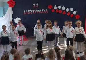 Dzieci ustawione w parach stoją na środku sceny. W tle dekoracja z okazji Święta Niepodległości