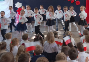 Dzieci trzymając się pod boki kołyszą się z nogi na nogę i śpiewają piosenkę o Krakowiaku. Młodsi koledzy siedzą na dywanie i oglądają występ "Słoneczek".