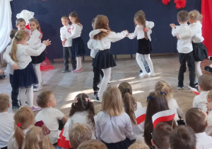 Dzieci tańczą Krakowiaka w parach poruszając się po kole. Młodsi koledzy siedzą na dywanie i oglądają występ "Słoneczek".
