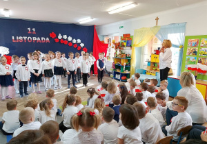 Pani dyrektor stoi obok tablicy tematycznej dziękując dzieciom za występ. Obok siedzą przedszkolaki z innych grup i Pani Małgosia.