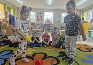 Klara i Franek stoją w kolorowych okręgach i rzucają kostkami do gry. Za nimi na dywanie siedzą dzieci.