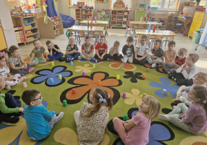 Dzieci siedzą w kole na dywanie. W środku koła widać rozłożone różnokolorowe plastikowe kubeczki.