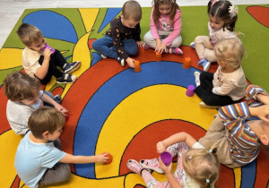 Dzieci siedzą w kole na dywanie podczas zabawy "Kubeczki do góry". W ręku każde dziecko trzyma kubeczek w różnym kolorze.