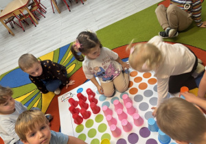 Na dywanie rozłożona jest mata do kodowania, na której znajduje się kolorwe kropki. Dzieci układają na poszczególnych polach kubeczki z adekwatnym kolorem do koloru kółka na macie.