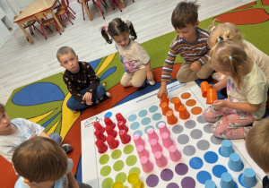 Leon, Kornelia i Maja układają pomarańczowe kubeczki na polach z pomarańczowymi kółeczkami. Reszta dzieci siedzi wokół maty.