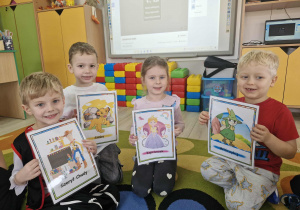 Oluś, Marcel, Ola i Karol siedzą na dywanie i trzymają w dłoniach plansze przedstawiające bohaterów bajek.