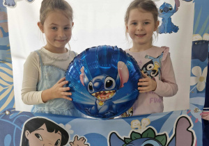 Oliwia i Ola pozują do zdjęcia przy foto - budce. Dziewczynki trzymają w dłoniach balon z wizerunkiem Sticha.