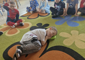 Dzieci siedzą w kole na dywanie. Na środku dywanu Marcel leży z poduszką pod głową udając, że śpi.