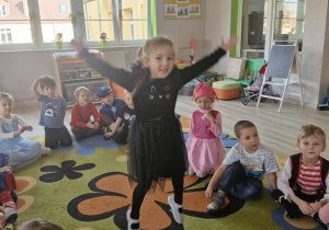 Zuzia skacze z rozłożonymi na boki rękami. W tle widać siedzące na dywanie dzieci.