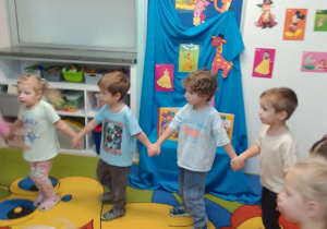 Dzieci poruszają się w kole przy muzyce. Z tyłu dekoracja z napisem "Dzień postaci z bajek", postacie z bajek z papieru i książki z bajkami.