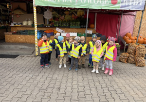Grupka dzieci w kamizelkach odblaskowych przed warzywniakiem.