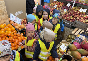 Dzieci w warzywniaku na tle owoców.