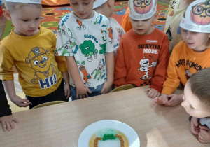 Grupka dzieci obserwująca doświadczenie z kolorowymi cukierkami ułożonymi w kształt dyni podlanych ciepłą wodą.
