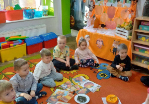 Grupka dzieci na dywanie z opaskami na głowie z sylwetą dyni obserwuje rozkrojoną dynię.