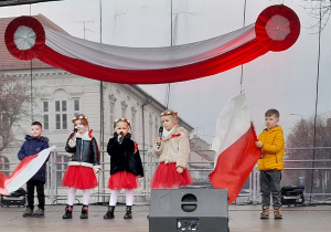 Pięcioro dzieci stoi na scenie na Placu Piłsudskiego. Trzy dziewczynki śpiewają piosenkę patriotyczną, a chłopcy poruszają biało-czerwonymi flagami.