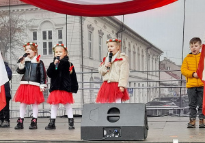 Dzieci śpiewają na scenie piosenkę "Symbole narodowe". Trzy dziewczynki stoją w środku, a po bokach chłopcy trzymają biało-czerwone flagi.