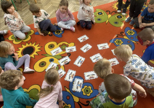 Dzieci siedzą na dywanie i zgadują do jakiej bajki należą atrybuty leżące na dywanie, przedstawione na obrazkach.