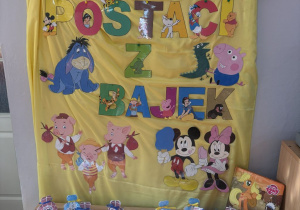 Dekoracja z napisem Dzień Postaci z Bajek wraz z postaciami z bajek, a pod dekoracją upominki w torebkach z logo postaci z bajek.