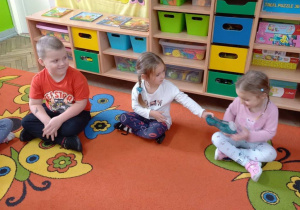 Felicja podaje Wiktorii pantofelek "Kopciuszka" w zabawie gorący pantofelek. Dzieci siedzą na dywanie.