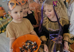 Szymuś trzyma w dłoniach dyniową miskę pełną smakołyków, a Lilianka torebkę z cukierkami. Za nimi widać inne dzieci z grupy