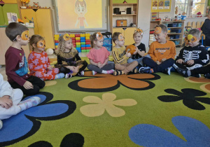 Dzieci siedzą w kole na dywanie i podają sobie dynię z rąk do rąk