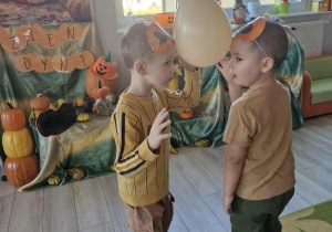 Marcel i Szymek stoją trzymając między głowami pomarańczowy balon. W tle widać dekorację przygotowaną z okazji Dnia Dyni