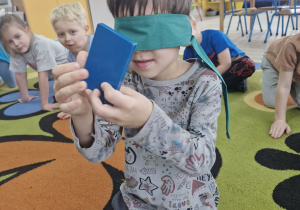 Wojtuś siedzi na dywanie. Chłopiec ma oczy zasłonięte szarfą, a w dłoniach trzyma niebieski prostokąt.