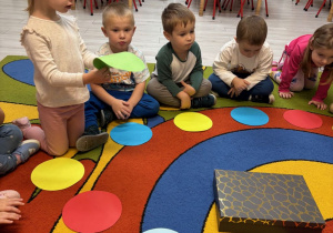 Dzieci siedzą w kole na dywanie z kolorowymi kółeczkami.