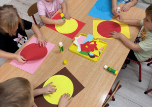 Dzieci siedzą przy stoliczkach i wykonują pracę plastyczną.