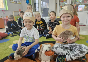 Ignaś i Wojtuś siedzą na dywanie w jeżykowych opaskach na głowie. Chłopcy trzymają w dłoniach maskotki jeżyków.