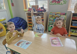 Marcel, Alicja i Weronika siedzą przy stoliku. Przed dziećmi leżą ułożone z części obrazki z wizerunkami jeży.