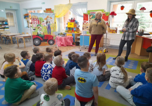 Dzieci siedzą na dywanie, a przed nimi stoi Pani Agata i jej córka. Panie witają przedszkolaków na warsztatach rolniczych. W tle banery z motywem wiejskim.