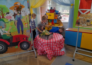 Czterech chłopców szuka jajek ukrytych w sali. Obok znajduje się stolik ze skrzynką z warzywami, banery o tematyce wiejskiej, kącik domowy.