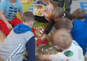 Grupka dzieci siedzi na podłodze i bawi się zagrodą wiejską, inne dzieci oglądają książki na dywanie.