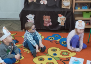 Dzieci oglądają obrazki i rozpoznają misie z różnych bajek. W tle dekoracja z napisem Dzień Misia i postaciami misiów.