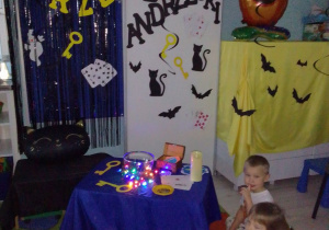 Dzieci siedzą na dywanie, w tle dekoracja z okazji Andrzejek: napis, sylwety czarnych kotów, duszków, kart, kluczy.