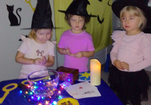Maja, Laura i Emilia w halloweenowych czapkach po wypowiedzeniu zaklęcia wrzucają do miski z wodą grosik na szczęście. Wokół inne "Pszczółki" obserwują ich działanie. Z tyłu dekoracja: napis Andrzejki, karty do gry, klucze, papierowe duszki.