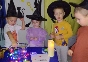 Szymon, Hania, Olaf i Michał wrzucają monety do wody do szklanej podświetlonej miski. Z tyłu andrzejkowa dekoracja.