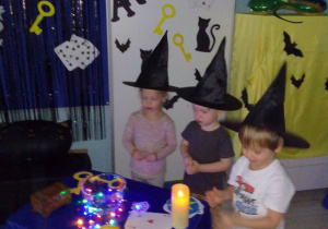 Ala, Olek i Nikoś w czarnych kapeluszach wrzucają do miski z wodą grosik na szczęście. Z tyłu dekoracja: napis Andrzejki, karty do gry, klucze, papierowe duszki.