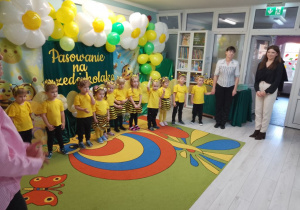 Dzieci w strojach pszczółek stoją na scenie. Za nimi dekoracja w stylu "Pszczółki na łące".