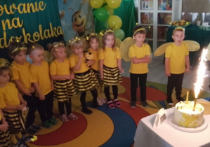 Dzieci w strojach pszczółek stoją w półkolu na dywanie, a przed nimi na stoliczku znajduje się tort z odpalonymi racami.
